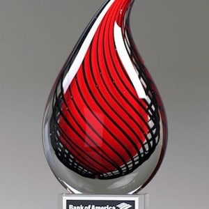 May include: A clear glass award shaped like a teardrop, with red and black striped design inside. The award sits on a clear base with a black and white nameplate that reads "Bank of America, Daniel Kariamis, Volunteer of the Year Award 2020."