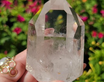 May include: A clear quartz crystal point with a double terminated point. The crystal is clear and has a slight cloudy appearance. The crystal is sitting on a hand.
