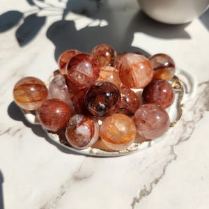 Op de afbeelding: Een verzameling kleine, ronde, roodbruine kristallen bollen, gepresententeerd in een wit schaaltje met een gouden rand. De bollen hebben interne patronen en kleurvariaties, variërend van helder tot donkerbruin. Het schaaltje staat op een wit marmeren oppervlak.