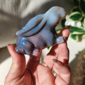 May include: A blue and purple agate bunny figurine. The bunny is carved from a translucent stone, with the ears and back in a lighter blue hue. The body has a darker purple and blue pattern. The figurine is in a crouching position, with its ears perked up.