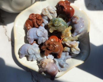 May include: A selection of small, carved stone elephant figurines in shades of red, blue, and green. The elephants are presented in a white, leaf-shaped dish. The figurines are displayed on a white surface with shadows.