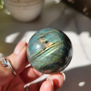 Peut inclure: Une sphère de labradorite polie et irisée. La pierre précieuse présente un mélange de bleus, de verts et de jaunes. La sphère est tenue dans une main, mettant en valeur sa surface lisse et réfléchissante. La sphère est une pierre naturelle.