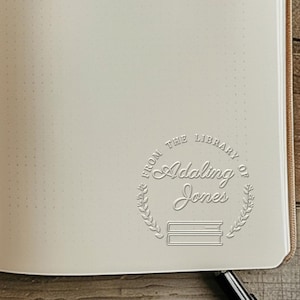 May include: Open notebook with dot grid pages. A circular stamp is embossed on the page, reading "FROM THE LIBRARY OF Adaling Jones" with a laurel wreath and two stacked books. The notebook is open to a blank page.