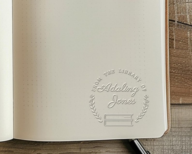 May include: Open notebook with dot grid pages. A circular stamp is embossed on the page, reading "FROM THE LIBRARY OF Adaling Jones" with a laurel wreath and two stacked books. The notebook is open to a blank page.