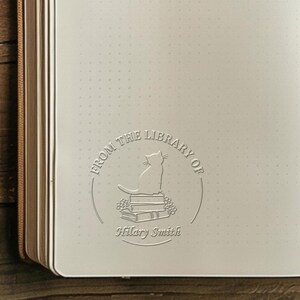 May include: Open journal with a debossed design featuring a cat sitting atop books, surrounded by the text "FROM THE LIBRARY OF" and the name "Hilary Smith". The journal has a dot grid pattern and a brown spine.