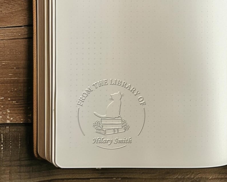 May include: Open journal with a debossed design featuring a cat sitting atop books, surrounded by the text "FROM THE LIBRARY OF" and the name "Hilary Smith". The journal has a dot grid pattern and a brown spine.