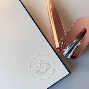 May include: A white book with a blue border and embossed text that reads "FROM THE LIBRARY OF ADALINE JONES" with a gift and book design. A rose gold book embosser is in the upper right corner.