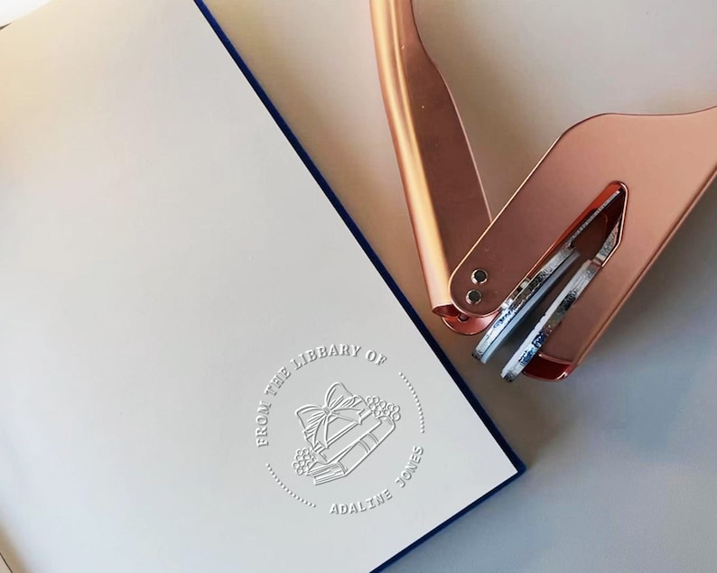 May include: A white book with a blue border and embossed text that reads "FROM THE LIBRARY OF ADALINE JONES" with a gift and book design. A rose gold book embosser is in the upper right corner.