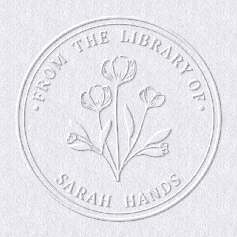 May include: A white embossed circular design with the text "FROM THE LIBRARY OF" at the top and "SARAH HANDS" at the bottom. The center features a floral illustration of three stylized flowers.