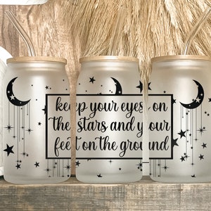 May include: Three frosted glass tumblers with bamboo lids and glass straws. Each tumbler features a black crescent moon and star design, with the quote "keep your eyes on the stars and your feet on the ground."