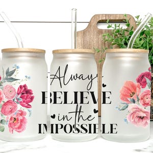Puede incluir: Tres vasos de vidrio esmerilado con forma de lata, con tapas de bambú y pajitas transparentes. Cada vaso presenta un diseño floral en tonos rosas y rojos. El vaso central tiene el texto "Always BELIEVE in the IMPOSSIBLE".