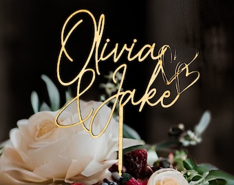 May include: Gold wedding cake topper with the names Olivia & Jake written in cursive script with two hearts.