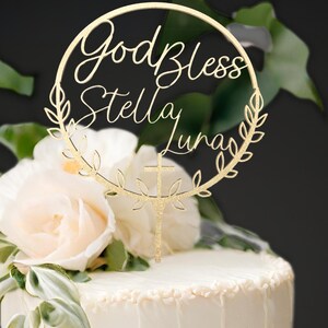 K&ouml;nnte beinhalten: Goldener Glitzer-Kuchentopper mit einem Kranz- und Kreuzdesign. Der Topper sagt "God Bless Stella Luna".