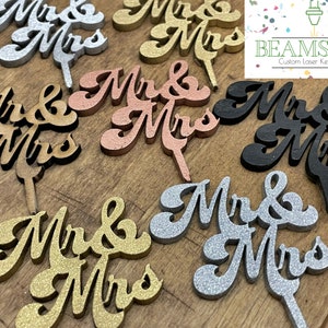 K&ouml;nnte beinhalten: Sortiment aus Holz "Mr & Mrs" Tortendekorationen in verschiedenen Farben und Ausf&uuml;hrungen, darunter Naturholz, Goldglitter, Silberglitter, Schwarz und Ros&eacute;gold. Die Dekorationen sind lasergeschnitten und haben eine kursive Schrift.