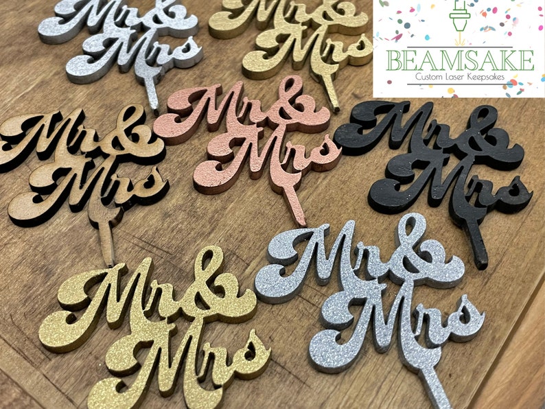 K&ouml;nnte beinhalten: Sortiment aus Holz "Mr & Mrs" Tortendekorationen in verschiedenen Farben und Ausf&uuml;hrungen, darunter Naturholz, Goldglitter, Silberglitter, Schwarz und Ros&eacute;gold. Die Dekorationen sind lasergeschnitten und haben eine kursive Schrift.