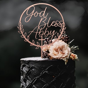 K&ouml;nnte beinhalten: Ein schwarzer Kuchen mit einer Rose und Beeren darauf. Ein Holzaufsatz mit den Worten "God Bless Abby" und einem Kreuz befindet sich oben auf dem Kuchen.