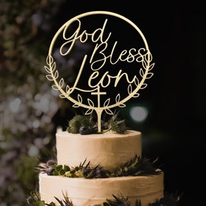 K&ouml;nnte beinhalten: Goldener Tortendekor mit einem Kranz und den Worten "God Bless Leon" in einer kursiven Schrift. Ein Kreuz befindet sich am unteren Ende des Kranzes.
