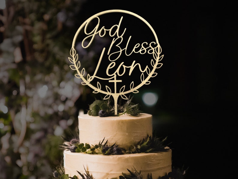K&ouml;nnte beinhalten: Goldener Tortendekor mit einem Kranz und den Worten "God Bless Leon" in einer kursiven Schrift. Ein Kreuz befindet sich am unteren Ende des Kranzes.