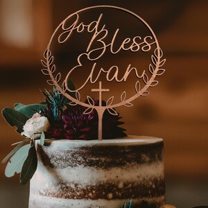 K&ouml;nnte beinhalten: Ein h&ouml;lzerner Tortendekor mit einem runden Design und den Worten "God Bless Evan" in Kursivschrift. Der Dekor ist mit einem Kreuz und Blattwerk verziert. Der Dekor befindet sich auf einer wei&szlig;en und braunen mehrst&ouml;ckigen Torte mit Blumen.