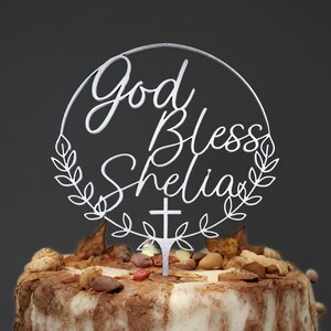 K&ouml;nnte beinhalten: Wei&szlig;er Tortendekor in Kreisform mit dem Text "God Bless Shelia" mit einem Kreuz und Bl&auml;ttern.