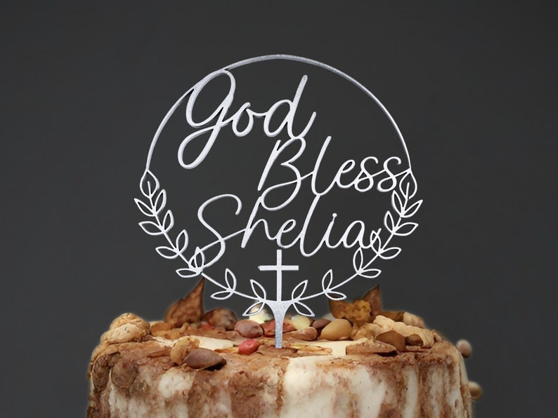 K&ouml;nnte beinhalten: Wei&szlig;er Tortendekor in Kreisform mit dem Text "God Bless Shelia" mit einem Kreuz und Bl&auml;ttern.
