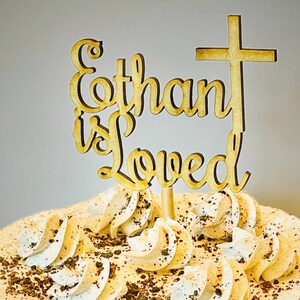 K&ouml;nnte beinhalten: Ein h&ouml;lzerner Kuchentopper mit den Worten "Ethan is Loved" und einem Kreuz oben drauf. Der Topper befindet sich auf einem Kuchen mit wei&szlig;er Zuckerglasur und Schokoladenstreuseln.