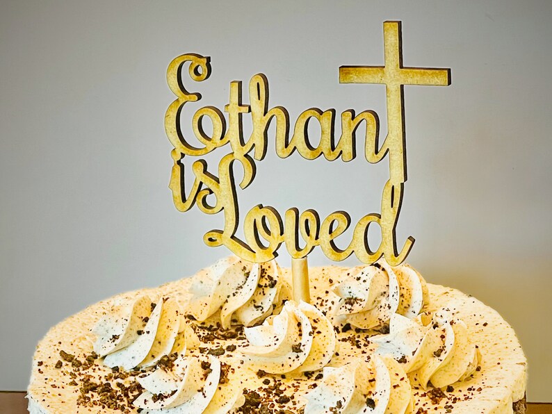 K&ouml;nnte beinhalten: Ein h&ouml;lzerner Kuchentopper mit den Worten "Ethan is Loved" und einem Kreuz oben drauf. Der Topper befindet sich auf einem Kuchen mit wei&szlig;er Zuckerglasur und Schokoladenstreuseln.