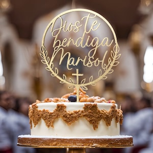 Könnte beinhalten: Ein weißer Kuchen mit einem goldenen Topper, auf dem "Dios te bendiga Marisol" und ein kleines goldenes Kreuz stehen. Der Kuchen ist mit Nüssen und Blaubeeren verziert.