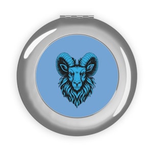 May include: A silver compact mirror with a blue ram illustration. The ram has large curved horns and a detailed design. The background of the mirror is light blue, and the compact has a shiny, metallic finish.