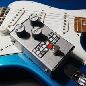 Könnte beinhalten: Ein silbernes Gitarreneffektpedal aus Metall mit drei schwarzen Drehreglern, beschriftet mit VOL, GAIN und TONE. Die Frontplatte trägt den Text FUZZ IKOL FX TITAN und S/N: 04-0226, mit einer leuchtenden roten LED neben einem Metallfußschalter.