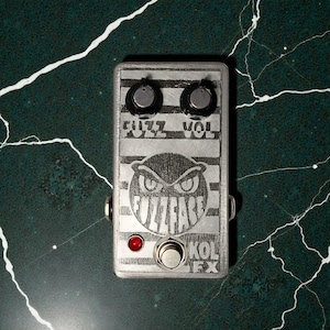 May include: Silver Fuzz Face guitar effects pedal with the text "FUZZ" and "VOL" above two black knobs. The pedal features an owl graphic and the text "FUZZFACE" and "KOL FX". A red light and a silver foot switch are also visible.