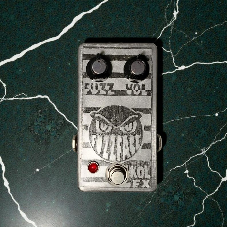 Pu&ograve; includere: Pedale per effetti per chitarra Fuzz Face argentato con le scritte "FUZZ" e "VOL" sopra due manopole nere. Il pedale presenta una grafica a gufo e le scritte "FUZZFACE" e "KOL FX". Sono visibili anche una luce rossa e un interruttore a pedale argentato.