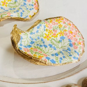 May include: Two gold-rimmed oyster shell dishes with a floral pattern in shades of pink, blue, and yellow.
