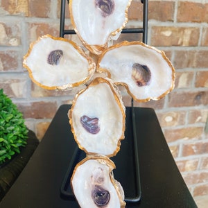 May include: A gold-trimmed oyster shell cross made of five oyster shells. The shells are arranged in a cross shape and are attached to a black metal stand.