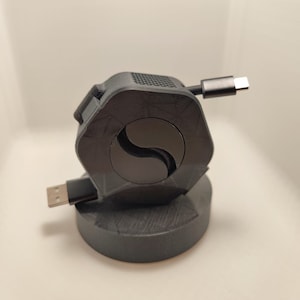 Könnte beinhalten: Ein schwarzer Kabelorganizer mit sechseckiger Basis und runder Oberseite. Das Kabel ist um die Oberseite gewickelt und die Enden sind in die Basis gesteckt.