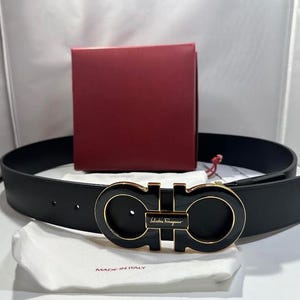 May include: A black leather belt with a gold-toned buckle featuring the Salvatore Ferragamo logo. The buckle is a double Gancini design. The belt is accompanied by a red box and a white dust bag with the words "Made in Italy" printed on it.