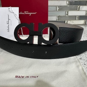May include: A black leather belt with a red and black interlocking logo buckle. The buckle has the text "Salvatore Ferragamo" on it. The belt is made in Italy.