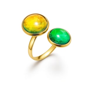 May include: A gold-toned open ring featuring two circular cabochon settings. The larger setting contains a yellow and green gradient stone, while the smaller setting holds a solid, vibrant green stone. The band is smooth and polished gold-tone metal.