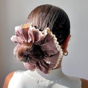 May include: A red and black checkered scrunchie with a cream-colored lace trim, worn in a bun. The scrunchie is made of a soft fabric and adds a touch of vintage flair to any hairstyle.