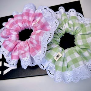 May include: Two gingham scrunchies with white lace trim. The scrunchies are pink and green. The green scrunchie has a small tag that says "Jansue".
