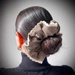 May include: A brown and white gingham scrunchie with a white lace trim, worn in a bun. The scrunchie is made of fabric and has a small tag. The person is wearing a black turtleneck.