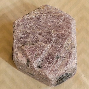 May include: A large, rough, pink ruby crystal with a hexagonal shape. The crystal has a natural, unpolished finish.