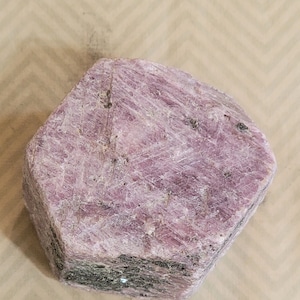 May include: A large, rough, pink ruby crystal with a hexagonal shape. The crystal has a natural, unpolished finish and is a beautiful example of this gemstone.