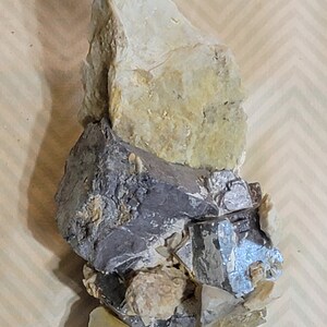 May include: A cluster of gray and silver crystals embedded in a light brown rock. The crystals have a metallic sheen and are arranged in a random pattern.
