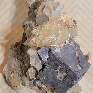 May include: A dark gray, cube-shaped mineral crystal sits on a bed of light gray and brown rock. The crystal is surrounded by smaller, clear, quartz crystals.