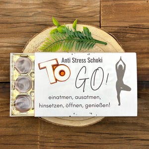 Könnte beinhalten: Eine Schachtel Pralinen mit dem Text "Anti Stress Schoki TO GO!" und einer Yoga-Pose-Silhouette. Die Schachtel enthält mehrere runde, silberfarbene Pralinen. Das Bild ist auf einem Holzgrund mit einem grünen Farn angeordnet.