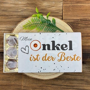 Peut inclure: Une boîte cadeau blanche avec le texte allemand "Mein Onkel ist der Beste" (Mon oncle est le meilleur) en lettres noires et dorées. La boîte contient des chocolats. La boîte est sur une surface en bois.