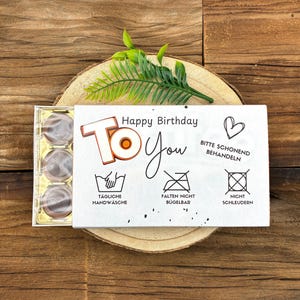 Puede incluir: Una caja de regalo con seis bombones, presentada sobre una rodaja de madera. La caja tiene una tarjeta blanca con el texto "Happy Birthday To You" y símbolos de cuidado. Una ramita verde decora la caja.