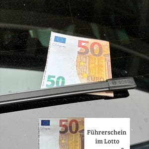 Könnte beinhalten: Ein Fünfzig-Euro-Schein liegt auf dem Heckscheibenwischer eines Autos. Der Schein ist orange, gelb und grün, mit der Zahl 50 deutlich sichtbar. Der Wischer ist schwarz, und der Hintergrund ist eine dunkle, reflektierende Oberfläche.