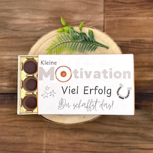 Peut inclure: Une boîte cadeau blanche avec l'inscription allemande "Kleine Motivation Viel Erfolg Du schaffst das !", contenant trois chocolats. La boîte est posée sur une tranche de bois, ornée d'une brindille de verdure.